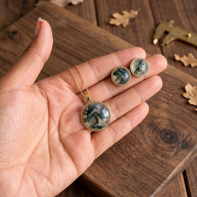 Elegant Moss Agate jewelry set displayed inside a luxury wooden jewelry box with soft pink velvet lining. The set includes a matching round Moss Agate pendant necklace and coordinating stud earrings, each featuring natural green moss-like inclusions within translucent white gemstones. The stones are bezel-set in gold-plated frames, creating a timeless and sophisticated look. This nature-inspired handmade jewelry set is perfect for weddings, bridesmaids, special occasions, and everyday wear,