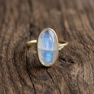 Könnte beinhalten: Ein goldener Ring mit einem ovalen, irisierenden Mondstein in Blau- und Weißtönen. Kleine Diamantakzente befinden sich auf beiden Seiten des Mondsteins. Der Ring ist vor einem rustikalen Holzgrund platziert.
