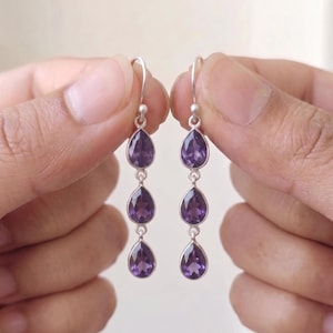 Könnte beinhalten: Ein Paar Ohrhänger mit jeweils drei vertikal angeordneten, tropfenförmigen violetten Amethyst-Edelsteinen in Silberfassung. Jeder Ohrring hängt an einem einfachen Silberhaken und zeigt insgesamt sechs facettierte violette Steine.