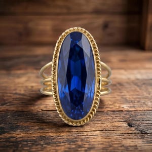 Pode incluir: Um anel de ouro com uma grande pedra preciosa azul-escura de corte oval, engastada numa borda de metal com design de corda torcida. A banda consiste em três fios de ouro finos e paralelos que se ligam ao engaste central da peça.