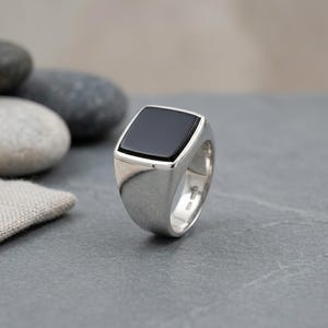 Könnte beinhalten: Ein silberner Ring mit einem rechteckigen schwarzen Onyxstein. Der Ring hat ein poliertes Finish und ein klassisches, minimalistisches Design. Der schwarze Stein ist in einem quadratischen Rahmen gefasst.