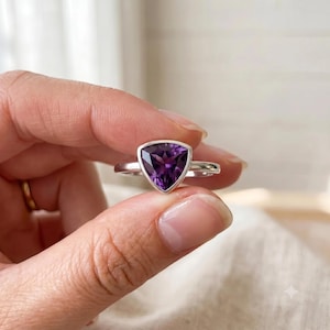 Könnte beinhalten: Ein Silberring mit einem facettierten, dreieckig geschliffenen violetten Amethyst, der in einer glatten Fassung sitzt. Der Ring verfügt über eine polierte Metallschiene, die den dreieckigen Stein hält und einen tiefen Violettton zeigt.