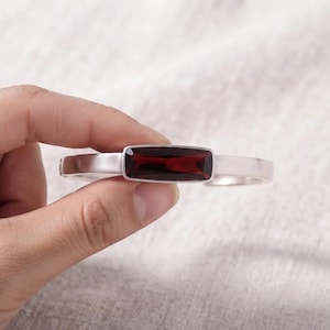 May include: A silver-toned bangle bracelet featuring a rectangular, deep red garnet gemstone set in the center. The metal band has a smooth, matte finish and a hinged design, showcasing a clean, horizontal orientation of the dark red stone.