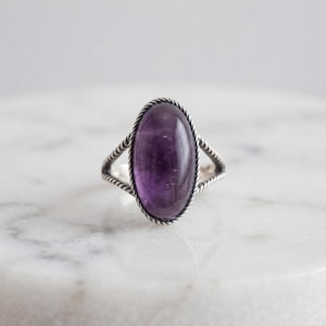 Könnte beinhalten: Ein Silberring mit einem großen, ovalen, violetten Amethyst-Edelstein in einer glatten Fassung. Die Ringschiene ist mit einer gedrehten, seilartigen Textur versehen, die sich in zwei Stränge teilt, während sie mit der zentralen Fassung verbunden ist.