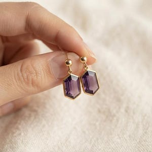 Könnte beinhalten: Ein Paar goldene Ohrhänger mit hexagonal geschliffenen violetten Amethyst-Edelsteinen. Jeder Ohrring hängt an einem kleinen goldenen Kugelstecker und zeigt einen facettierten, durchscheinenden violetten Stein in einem polierten Goldrahmen.