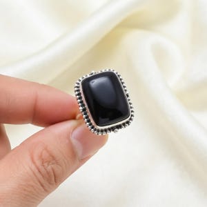 Könnte beinhalten: Ein Silberring mit einem großen, rechteckigen, polierten schwarzen Stein, der in einen Rahmen mit perlenartigem, strukturiertem Rand gefasst ist. Das Metallband hat eine helle, reflektierende Oberfläche und der Stein ist glatt und glänzend.
