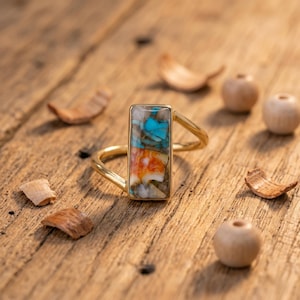 Peut inclure: Une bague dorée avec une incrustation rectangulaire composée de fragments de pierre turquoise, orange et blanche. L'anneau est fin et métallique, présentant une finition polie qui contraste avec la pièce centrale en pierre texturée et multicolore.