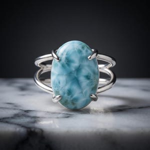 Könnte beinhalten: Ein Silberring mit einem großen, ovalen, hellblauen Larimar-Edelstein mit weißen Marmormustern. Der Stein wird von vier Krappen gehalten und die Ringschiene besteht aus zwei parallelen Silberdrähten, die sich zu beiden Seiten teilen.