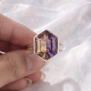May include: A silver ring featuring a large, faceted, hexagonal-cut gemstone. The stone displays a distinct color gradient, transitioning from a warm golden-yellow on one side to a deep, vibrant purple on the other, set in a simple metal band.