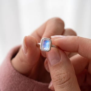 May include: A silver ring featuring a rectangular, emerald-cut moonstone set in a smooth bezel setting. The gemstone displays a translucent, iridescent blue glow within its faceted surface, mounted on a simple, polished silver band.