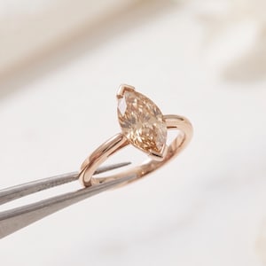 Könnte beinhalten: Ring mit Diamanten im Marquise-Schliff und einem Roségoldband. Der Diamant hat eine hellchampagnerfarbene Farbe und wird von einer Vier-Zacken-Fassung gehalten. Der Ring wird von einer Pinzette vor weißem Hintergrund gehalten.