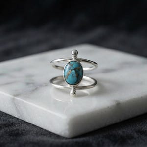 Könnte beinhalten: Ein Silberring mit einem zentralen ovalen Türkisstein mit brauner Matrix-Maserung. Die Ringschiene besteht aus zwei parallelen Silberdrähten, die am Stein zusammenlaufen und durch kleine Silberkugeln an der Fassung akzentuiert werden.