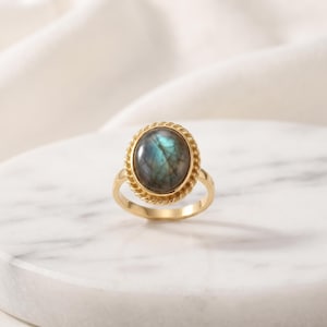 Könnte beinhalten: Ein goldener Ring mit einem zentralen, ovalen Labradorit-Edelstein in bläulichen und graulichen irisierenden Tönen. Der Stein ist in eine gedrehte, seilartige Metallfassung eingefasst, die auf einer glatten, polierten Goldschiene sitzt.