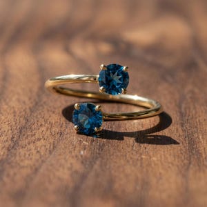 Könnte beinhalten: Ein goldfarbener Ring mit zwei runden, tiefblauen Edelsteinen. Das Band des Rings verläuft geschwungen und hält die Steine in einem einzigartigen, modernen Design. Der Ring ist auf einer warmen Holzoberfläche platziert, was seine Farbe und sein Funkeln hervorhebt.