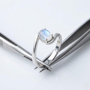 Könnte beinhalten: Ein silberner Ring mit einem hellblauen, ovalen Edelstein. Der Ring hat ein einzigartiges, fließendes Design. Der Ring wird von einer silbernen Pinzette vor weißem Hintergrund gehalten. Der Edelstein scheint ein Mondstein zu sein.