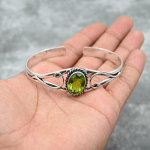 Könnte beinhalten: Ein silberfarbenes Armreif-Armband mit einem ovalen, hellgrünen Edelstein in der Mitte. Der Edelstein ist von einem gedrehten Seildesign umgeben. Dekorative Wirbel zieren beide Seiten. Schmuck.