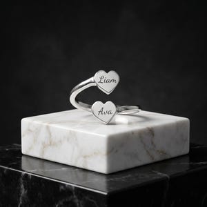 Könnte beinhalten: Ein silberfarbener Wickelring mit zwei herzförmigen Enden. Jedes Herz ist mit einem Namen in Schreibschrift graviert, wobei das obere Herz den Namen Liam und das untere Herz den Namen Ava zeigt. Der Ring hat eine polierte Oberfläche.