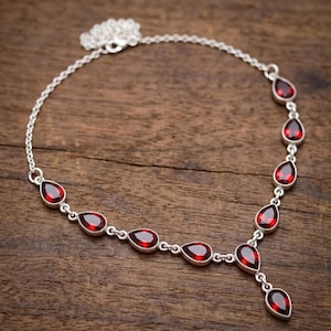 Pode incluir: Um colar de corrente prateada com várias pedras preciosas vermelho-escuras em forma de pera, engastadas em molduras de metal. O colar apresenta um design de corrente de elos delicados com um elemento central pendente em forma de gota.