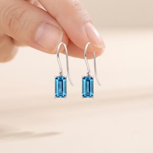 Könnte beinhalten: Ein Paar silberne Ohrhänger mit rechteckigen, smaragdgeschliffenen blauen Edelsteinen. Jeder Ohrring hängt an einem einfachen silbernen Haken und zeigt eine klare, leuchtend blaue Farbe sowie eine polierte Metallfassung mit vier Krappen.