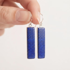 May include: A pair of rectangular drop earrings featuring deep blue lapis lazuli stones with gold-colored flecks, set in silver-toned metal frames with curved ear wires for a secure fit and a polished finish.