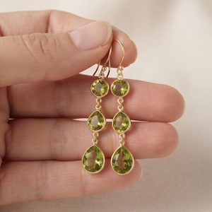 Könnte beinhalten: Ein Paar goldfarbene Ohrhänger mit jeweils drei facettierten, hellgrünen Edelsteinen im Peridot-Stil. Der obere Stein ist rund, während die mittleren und unteren Steine tropfenförmig sind und in Metallfassungen mit Hakenverschlüssen sitzen.