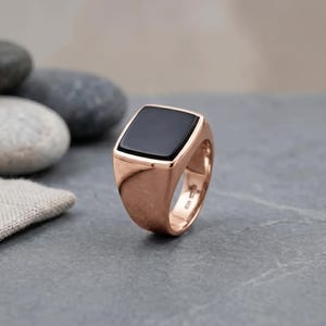 Könnte beinhalten: Ein Siegelring aus Roségold mit einer flachen, rechteckigen Einlage aus schwarzem Onyx. Die Ringschiene hat eine polierte metallische Oberfläche mit einer glatten, massiven Konstruktion und einem sichtbaren Stempel auf der Innenseite.