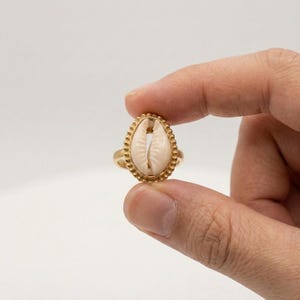 Könnte beinhalten: Ein goldfarbener Ring mit einem cremefarbenen Muschelschmuckstück. Die Muschel ist oval und in einen goldfarbenen, perlenbesetzten Rahmen gefasst. Der Ring wird zwischen zwei Fingern vor weißem Hintergrund gehalten, um das Design des Schmucks hervorzuheben.