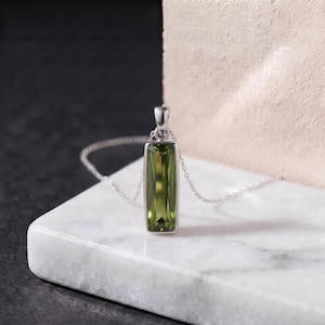 May include: A silver pendant necklace featuring a long, rectangular, faceted olive green gemstone set in a polished silver frame. The pendant hangs from a delicate silver chain, showcasing the deep green color and clear, geometric cut of the stone.