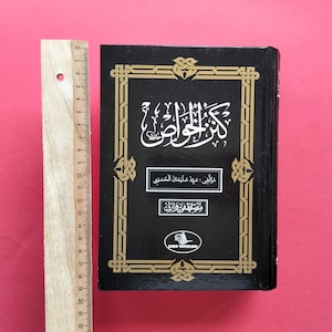 Op de afbeelding: Een zwart hardcover boek met gouden Arabische kalligrafie en decoratieve randpatronen op de voorkant. Een houten liniaal is verticaal naast het boek geplaatst en toont een hoogte van ongeveer 22 centimeter.
