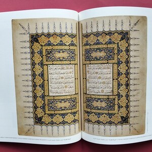 Könnte beinhalten: Ein aufgeschlagenes Buch mit zwei Seiten komplexer arabischer Kalligrafie, eingerahmt von kunstvollen Bordüren in Gold-, Dunkelblau- und Schwarztönen. Der Text ist in rechteckigen Feldern zentriert und von detaillierten Blumenmotiven umgeben.