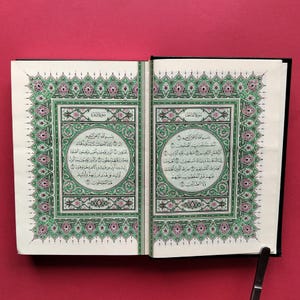 Peut inclure: Un livre ouvert avec deux pages présentant des bordures symétriques complexes dans des tons de vert, rose et noir. Le texte est écrit en calligraphie arabe, centré dans des cadres circulaires décoratifs sur chaque page, sur un fond rouge uni.