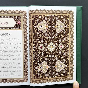 May include: An open hardback book with a dark green cover. The left page features Arabic text in a decorative frame, while the right page displays an intricate, symmetrical floral pattern in shades of brown, cream, and gold. The design is ornate.