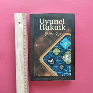 Peut inclure: Un livre noir intitulé Uyunel Hakaik avec une couverture sur le thème céleste comportant des formes géométriques et du texte. Une règle en bois est placée à côté du livre, montrant une hauteur d'environ 23 cm pour la couverture.