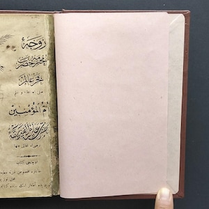 Może przedstawiać: Otwarta zabytkowa książka w brązowej oprawie z ilustracją dwóch palm na lewej stronie. Strona zawiera skomplikowaną czarną arabską kaligrafię i tekst, w tym rok 1322. Prawa strona to pusta, jasnoróżowa kartka.