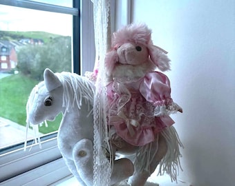 Könnte beinhalten: Ein weißes Plüsch-Karussellpferd mit weißer Wollmähne und Schweif, montiert auf einem rechteckigen Sockel. Ein rosa Plüschpudel in einem rosa Satinkleid mit weißer Spitzenverzierung sitzt auf dem Pferd. Ein weißes Spitzenband hängt von der Stange.