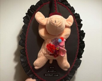 Könnte beinhalten: Ein Plüschschwein mit einem schwarzen Partyhut und einer roten Schleife, montiert auf einer ovalen schwarzen Platte mit schwarzem Rüschenrand und roter Verzierung. Das Schwein hat ein buntes Stoffbündel am Bauch und den Text SUS SCROPA DOMESTICUS.