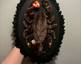 Könnte beinhalten: Ein braunes Plüsch-Skorpion-Spielzeug, montiert auf einer ovalen schwarzen Platte mit einem gerüschten schwarzen Stoffrand. Der Skorpion hat ein rotes herzförmiges Ty-Etikett an der Schere und kleine goldene Herzakzente an den Beinen. Das Wort BONES ist aufgedruckt.