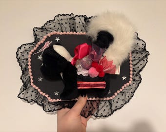 Könnte beinhalten: Eine schwarz-weiße Plüsch-Stinktierfigur auf einer schwarzen ovalen Platte. Die Platte ist mit rosa Pompons und schwarzem Tüll mit Punkten umrandet. Das Stinktier ist mit rosa und roten Bändern, Stoffblumen und kleinen silbernen Sternen verziert.