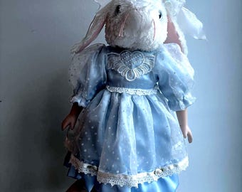 Könnte beinhalten: Eine Plüschhasenpuppe mit weißem Fell und langen Ohren, bekleidet mit einem blauen Satinkleid mit weißem, gepunktetem Organza-Überwurf. Das Kleid hat einen herzförmigen Spitzenkragen, Puffärmel und eine weiße Spitzenborte am Saum. Der Hase trägt weiße Schleifen auf dem Kopf und kleine weiße Schuhe.