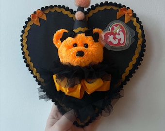 Könnte beinhalten: Ein orangefarbener Plüschbär mit schwarzen Augen und Nase, der einen schwarzen Partyhut mit einem pfirsichfarbenen Bommel, schwarze Tüllrüschen und ein gelbes Band trägt. Der Bär ist auf einer schwarzen herzförmigen Platte mit Goldrand und Ty-Etikett befestigt.