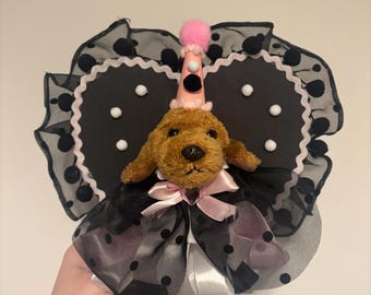 Könnte beinhalten: Ein brauner Plüsch-Hundekopf mit einem hellrosa Partyhut und einem rosa Bommel, zentriert auf einer schwarzen herzförmigen Unterlage mit weißen Punkten und einem gerüschten schwarzen Organzasaum. Eine hellrosa Schleife sitzt unter dem Kinn.