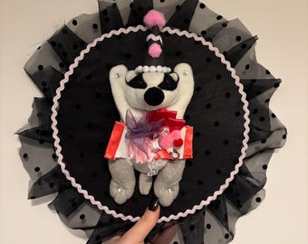 Könnte beinhalten: Eine kreisförmige Wanddekoration mit einer weißen und grauen Plüschtierfigur, die einen schwarzen Partyhut mit rosa Bommeln trägt. Die Figur ist auf einer schwarzen, gepunkteten Stoffscheibe mit schwarzem Tüllrand und rosa Besatz zentriert.