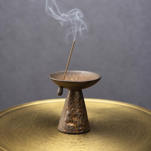 May include: A dark brown, textured ceramic incense burner with a wide, shallow bowl and a tapered, conical base. A single thin incense stick stands upright in the center of the bowl, emitting a visible trail of white smoke into the air.