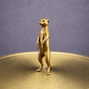 May include: A bronze meerkat figurine stands upright on a gold-colored surface. The meerkat is detailed with a textured finish, showcasing its alert posture and small, attentive eyes. The figurine is a decorative item.