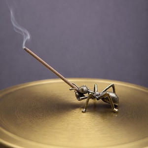 May include: A brass incense holder shaped like an ant, holding a lit incense stick. The ant is positioned on a gold-colored, round tray. Wisps of smoke rise from the incense stick against a dark background. The overall theme is home decor and relaxation.
