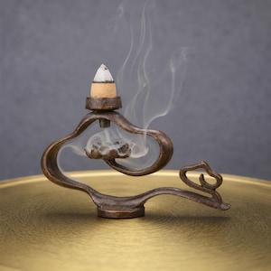 May include: A bronze-toned metal incense burner with a curved, fluid frame and a lotus-shaped base. A lit incense cone sits on top, releasing a thin trail of white smoke that flows downward through the center of the structure into the lotus.