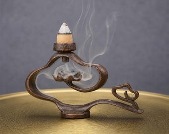 May include: A bronze-toned metal incense burner with a curved, fluid frame and a lotus-shaped base. A lit incense cone sits on top, releasing a thin trail of white smoke that flows downward through the centre of the structure into the lotus.
