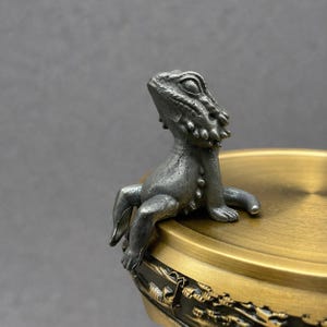 May include: A detailed, silver-colored metal lizard figurine perched on the edge of a brass-colored container. The lizard has textured skin and is looking upwards. The container has a gold finish and decorative detailing.