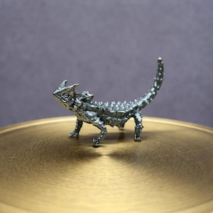 May include: A detailed silver-colored metal figurine of a thorny devil lizard. The lizard has a spiky body, a long tail, and is posed on a gold-colored, circular surface. The background is a muted purple.