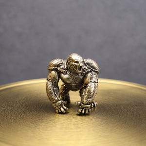 May include: A small, metallic bronze-colored figurine of a gorilla in a crouching, aggressive stance. The figure features intricate, mechanical-looking armor plating on its shoulders and arms, with a textured finish and dark, recessed detailing.